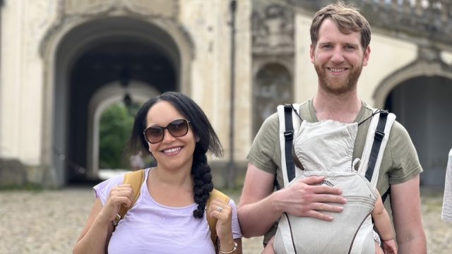 Family standing side-by-side. Wife, husband carrying baby in a baby carrier. In the background, is a large yellow castle located in Stuttgart, Germany,