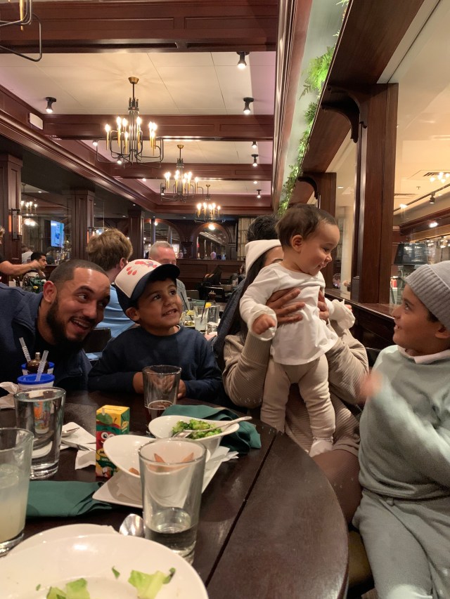 The family is gathered around the dinner table at a restaurant. There is a man, a woman, and three small children. They are all facing the small little girl who is held by the woman.