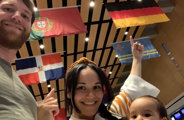 Husband, wife and newborn baby standing under two large flags representing Dominican Republic and Germany. The family is happy as they are moving abroad to Germany.