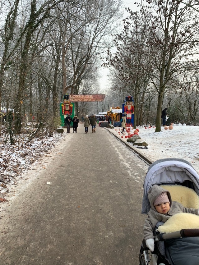 Small child in a stroller standing in front of the Christmas market entrance. There is some snow on the ground, a sign of winter.