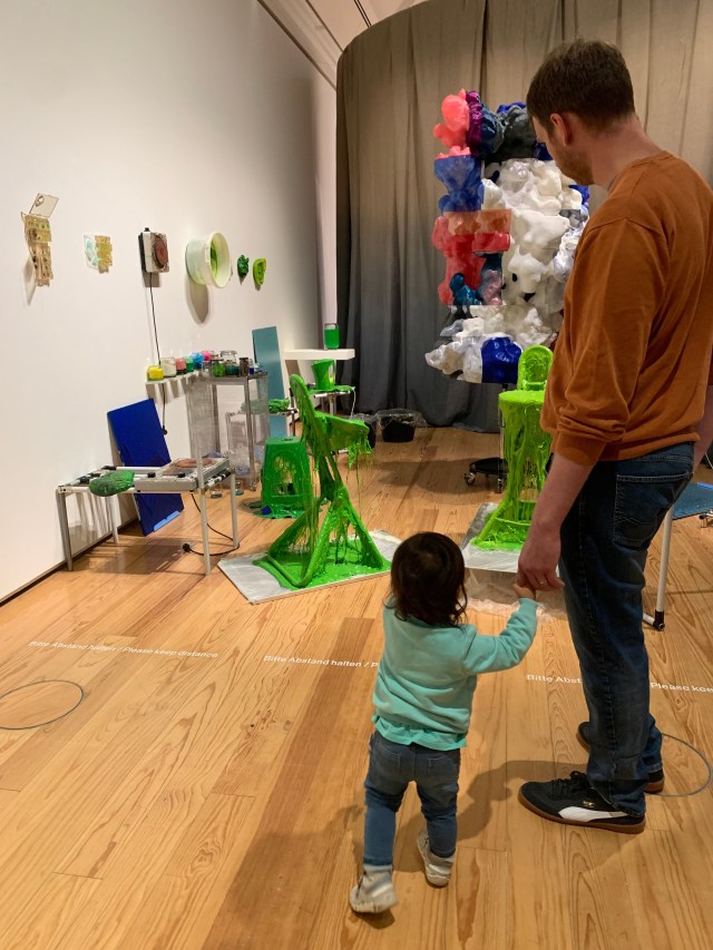 Small child standing next to her father holdings hands while admiring an art exhibition with their backs turned to us.