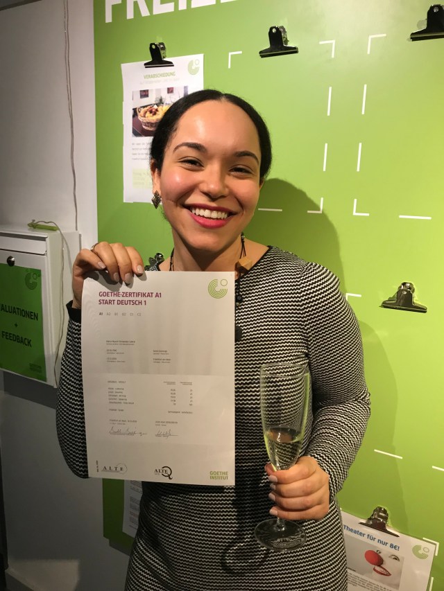 A young woman in her 30's proudly holding a language certificate from Goethe Institute. Celebrating with a glass of champagne achieving her A1 German language certificate.